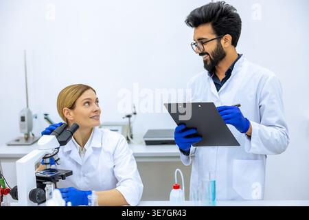 Lächelnder indischer Wissenschaftler, der Labormantel und Latexhandschuhe trägt, macht Notizen, nachdem er einen Beispieltest in der Nähe einer kaukasischen Kollegin mit Mikrosko gemacht hat... Stockfoto