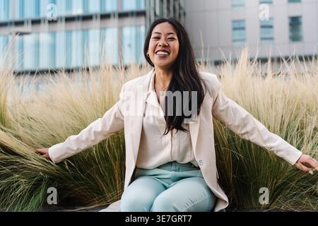 Fröhliche asiatische Geschäftsfrau, die auf Gras in der Nähe von Gebäuden auf der Stadtstraße sitzt Stockfoto