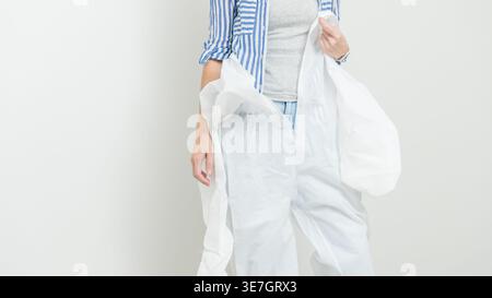 Frau in Schutzkleidung hält einen weißen Overall und trägt ein gestreiftes Hemd, bereit für die Renovierung in einem hellen Innenraum Stockfoto