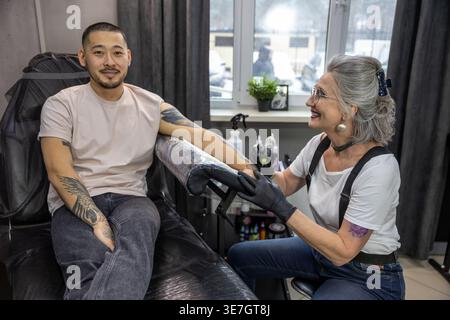 Lächelnder asiatischer Mann, der auf einem Stuhl sitzt und von einer kaukasischen Künstlerin mittleren Alters im Studio tätowiert wird Stockfoto