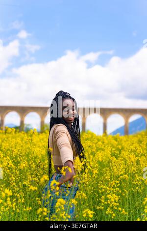 Lächelnde schwarze Frau mit Wendungen, die gelbe Blume berühren, steht auf dem Feld an sonnigem Tag Stockfoto