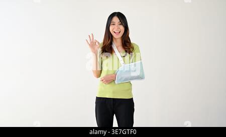 Lächelnde asiatische Frau, die einen Schlingenarm trägt und eine gute Handgeste vor grauem Hintergrund zeigt Stockfoto
