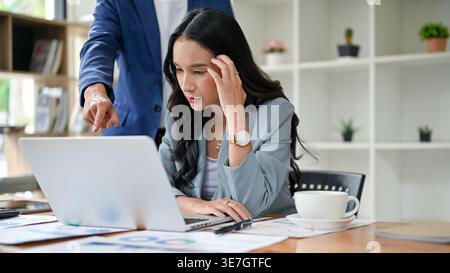 Gestresste asiatische Büroangestellte, die von ihrem männlichen Chef an ihrem Schreibtisch im Büro beschwert wurde Stockfoto