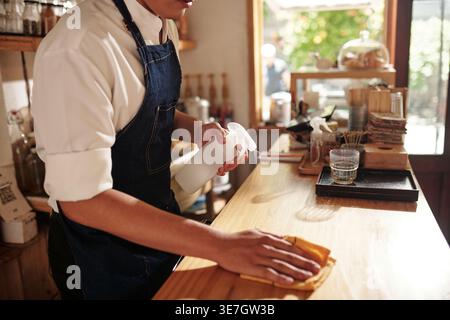Asiatische männliche Kellner Putztisch mit Teppich und Desinfektionsspray, während sie an der Theke im Café stehen Stockfoto
