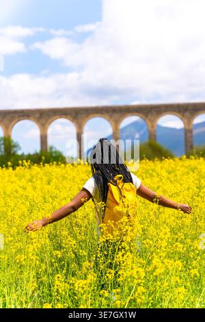 Schwarze Frau mit Wendungen, die Rucksack mit ausgestreckten Armen im Feld der gelben Blumen an sonnigem Tag trägt Stockfoto