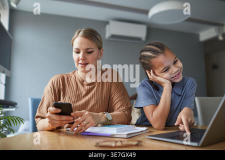 Schwarzes Mädchen mit Zöpfen mit Laptop und kaukasischer Mutter mit Smartphone, das zu Hause am Tisch in der Küche sitzt Stockfoto