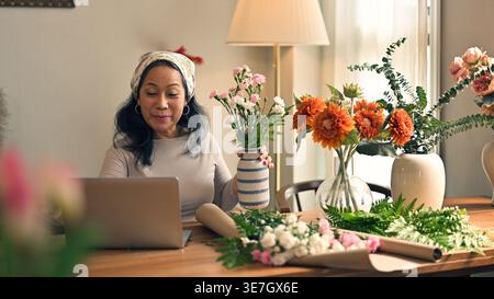 Lächelnde ältere asiatische Frau, die ein Bandana trägt, eine Vase mit Blumen hält und einen Laptop benutzt, während sie zu Hause mit Blumenarrangements am Tisch sitzt Stockfoto