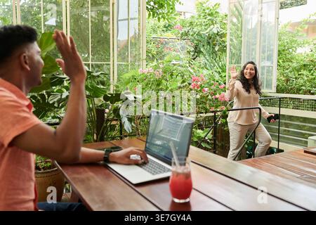 Ein indischer Mann sitzt am Tisch, arbeitet am Laptop und winkt der asiatischen Frau, die im Café zu ihm geht Stockfoto