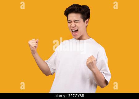Asiatischer Mann mit weißem Hemd, der im Studio mit geschlossenen Augen vor der orangefarbenen Wand schreit und seine Fäuste geballt Stockfoto