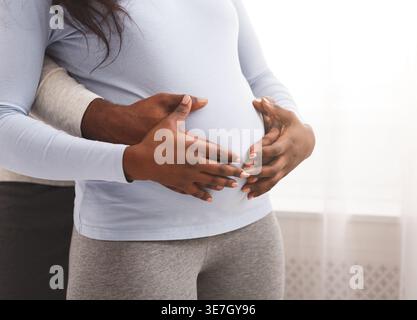 Zugeschnittenes Bild von Schwarzen Mann, der schwangere Frau von hinten umarmt und tagsüber im Schlafzimmer steht Stockfoto