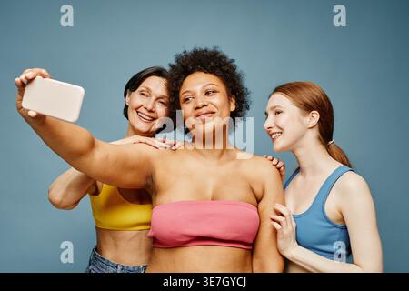 Gemischte Rassenfrau, die Selfie mit kaukasischen Freundinnen macht, die zusammen vor blauem Hintergrund im Studio stehen Stockfoto