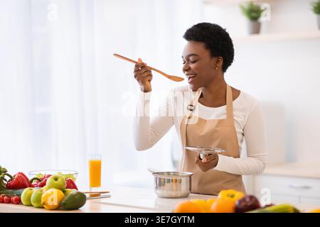 Schwarze Frau mit kurzen Haaren, die Schürze trägt, Kochen und Verkostung mit Holzlöffel, während sie zu Hause an der Küchentheke steht Stockfoto