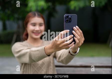 Thailändische Frau nimmt Selfie mit Smartphone auf, während sie tagsüber auf der Bank im Park sitzt Stockfoto
