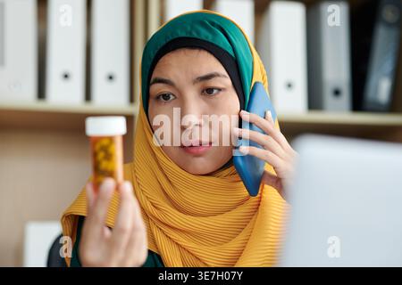 Eine Frau mit gemischter Rasse, die einen Hijab trägt und eine Medikamentenflasche hält, während sie am Tisch im Büro telefoniert Stockfoto