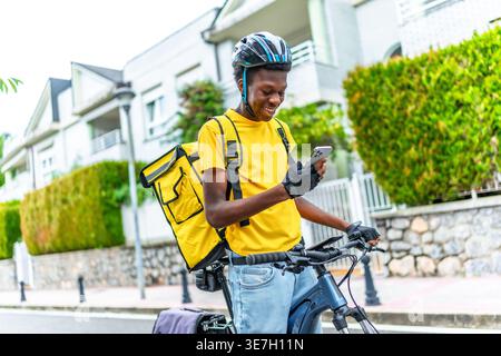 Schwarzer Mann, der Helm und Liefertasche trägt und das smLifestylephone benutzt, um den Kunden zu kontaktieren, während er tagsüber mit dem Fahrrad auf der Straße steht Stockfoto
