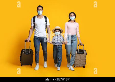 Arabische Familie trägt Gesichtsmasken, die Hände halten und ihr Gepäck ziehen, während sie im Studio vor gelbem Hintergrund läuft Stockfoto