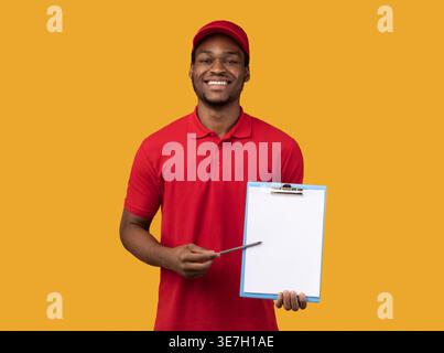 Der lächelnde Schwarze Mann trägt eine Mütze, die Stift und Klemmbrett mit weißem Papier hält und im Studio vor gelbem Hintergrund steht Stockfoto