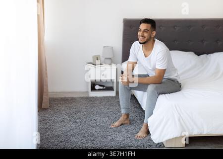 Ein arabischer Mann, der wegschaut, während er zu Hause auf dem Bett sitzt Stockfoto