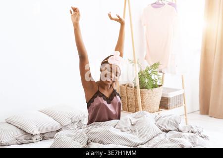 Schwarze Frau mit Augenmaske auf der Stirn, die Arme nach dem Aufwachen während sie zu Hause auf dem Bett sitzt Stockfoto