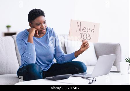 Schwarze Frau mit einem kurzen Afro-Schild, das ein Schild aus Pappe hält, mit Schreibtext, während sie Videoanrufe über einen Laptop im Wohnzimmer zu Hause hat Stockfoto