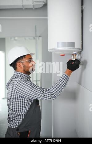 Indischer Klempner mit Uniform und Schutzhelm, der im Bad an der Heizung steht und arbeitet Stockfoto