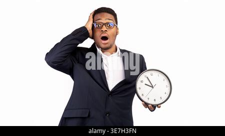 Schockierter schwarzer Geschäftsmann, der eine Wanduhr vor weißem Hintergrund hält Stockfoto