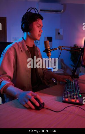 Asiatischer männlicher Online-Spiel-Streamer, der am Mikrofon spricht, während er Videospiele auf dem Computer spielt, der zu Hause auf dem Stuhl sitzt Stockfoto