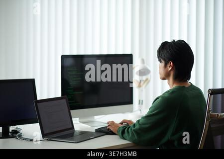 Asiatischer männlicher Programmierer, der Codes am Computer eingibt, während er mit Laptop im Büro am Tisch sitzt Stockfoto