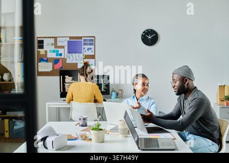 Schwarze männliche und Mixed Race weibliche Programmierer mit Brille sitzen am Schreibtisch und diskutieren Computerprogramme im Büro Stockfoto