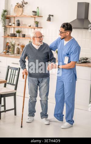 Lächelnder arabischer männlicher Betreuer in Brille, der einen älteren Mann im Stehen zu Hause ansieht Stockfoto
