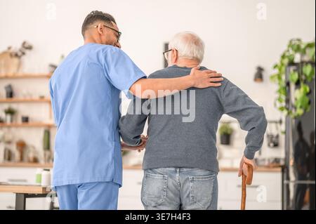 Lächelnder arabischer männlicher Betreuer in Brille, der einen älteren Mann im Stehen zu Hause ansieht Stockfoto