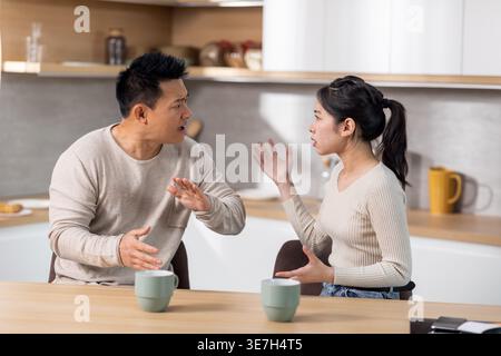 Wütende asiatische Frau streitet mit ihrem Mann, der sich anschrie und Gesten machte, während er am Tisch saß und in der Küche zu Hause Kaffee trank Stockfoto