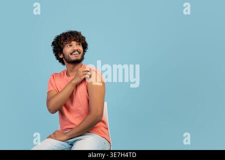 Bärtiger indischer Mann mit lockigem Haar, der einen geimpften Arm zeigt, während er auf Stuhl vor blauem Hintergrund im Studio sitzt Stockfoto