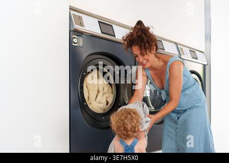 Hispanische Mutter lädt eine Waschmaschine mit Sohn, der ihr hilft, während er in der Wäscherei steht Stockfoto