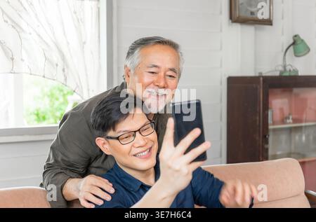 Asiatischer älterer Vater, der hinter einem Mann mittleren Alters steht und ein Smartphone benutzt, das auf der Couch sitzt, während beide im Wohnzimmer zu Hause Videoanrufe führen Stockfoto