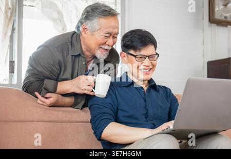 Asiatischer Mann mittleren Alters, der auf der Couch sitzt und einen Laptop benutzt, während ein älterer Vater eine Kaffeetasse hält, die daheim hinter ihm steht Stockfoto