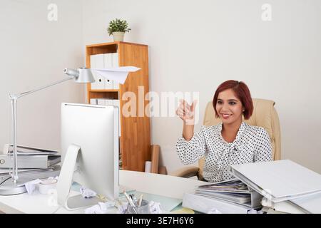 Indische Geschäftsfrau wirft Papierflugzeug und sitzt am Schreibtisch voll mit Dokumenten und zerknitterten Papieren im Büro Stockfoto