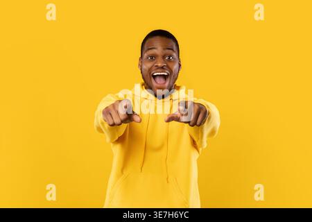 Der aufgeregte bärtige Schwarze Mann trägt einen Hoodie, der die Finger nach vorne zeigt, während er im Studio mit geöffnetem Mund vor gelbem Hintergrund steht Stockfoto
