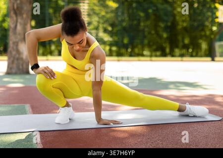 Schwarze Frau in Fitnesskleidung Stretching Bein tun seitliche Ausfallschritte Training auf Yogamatte im Sportpark an sonnigem Tag Stockfoto