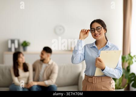Nahöstliche Psychologin mit Brille und Klemmbrett, während sie in der Klinik gegen Ehepaare steht Stockfoto