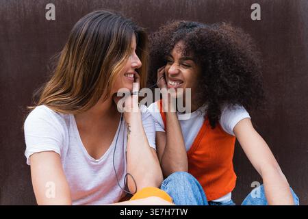 Hispanische Frau mit lockigem Haar, die Musik auf Ohrhörern hört, während sich kaukasische Freundin anschaut, während sie an der braunen Wand sitzt ... Stockfoto