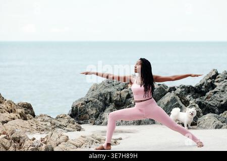 Schwarze Frau mit Dreadlocks im Fitness-Outfit, die Arme und Beine am Meer dehnt Stockfoto