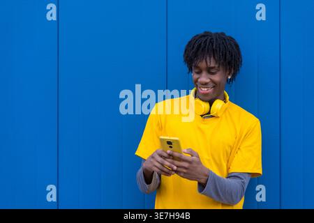 Ein lächelnder Schwarzer Mann mit Wendungen, der ein gelbes Hemd und Kopfhörer am Hals trägt und das Smartphone benutzt, während er auf der Straße an der blauen Wand steht Stockfoto