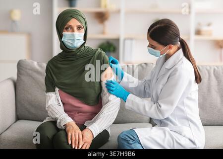 Nahost-Ärztin in Gesichtsmaske und Handschuhen, die Gipsverband auf den geimpften Arm einer muslimischen Frau mittleren Alters im Hijab während des Sitzens ... aufträgt Stockfoto