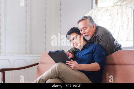 Asiatischer älterer Vater, der sich hinter seinem Sohn mittleren Alters mit einer Brille in der Hand eines Tablets auf der Couch zu Hause liegend Stockfoto