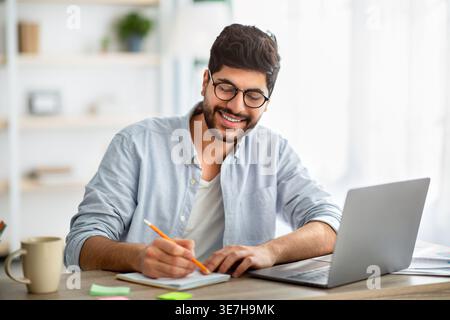 Lächelnder arabischer Mann mit Bart, der eine Brille trägt und Notizen macht, während er am Laptop arbeitet und zu Hause am Schreibtisch sitzt Stockfoto