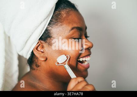 Große hispanische Frau trägt ein Badetuch um den Kopf und hat Spaß mit der Gesichtsrolle und lacht, während sie seitlich vor grauem Hintergrund steht ... Stockfoto