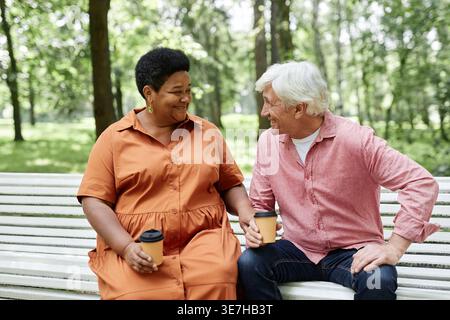 Schwarze Frau mittleren Alters, die einen Kaffee mit einem älteren kaukasier auf einer Holzbank im Park hat Stockfoto