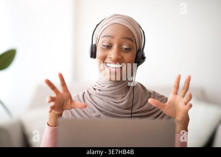 Schwarze muslimische Geschäftsfrau mit Headset, die Videoanrufe auf einem Laptop am Tisch zu Hause im Wohnzimmer führt Stockfoto
