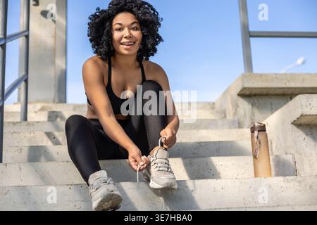 Schwarze Frau mit lockigen Haaren, die an sonnigen Tagen im Stadion auf Treppen sitzt und ein schwarzes Oberteil und Leggings trägt, die ihre Turnschuhe binden Stockfoto